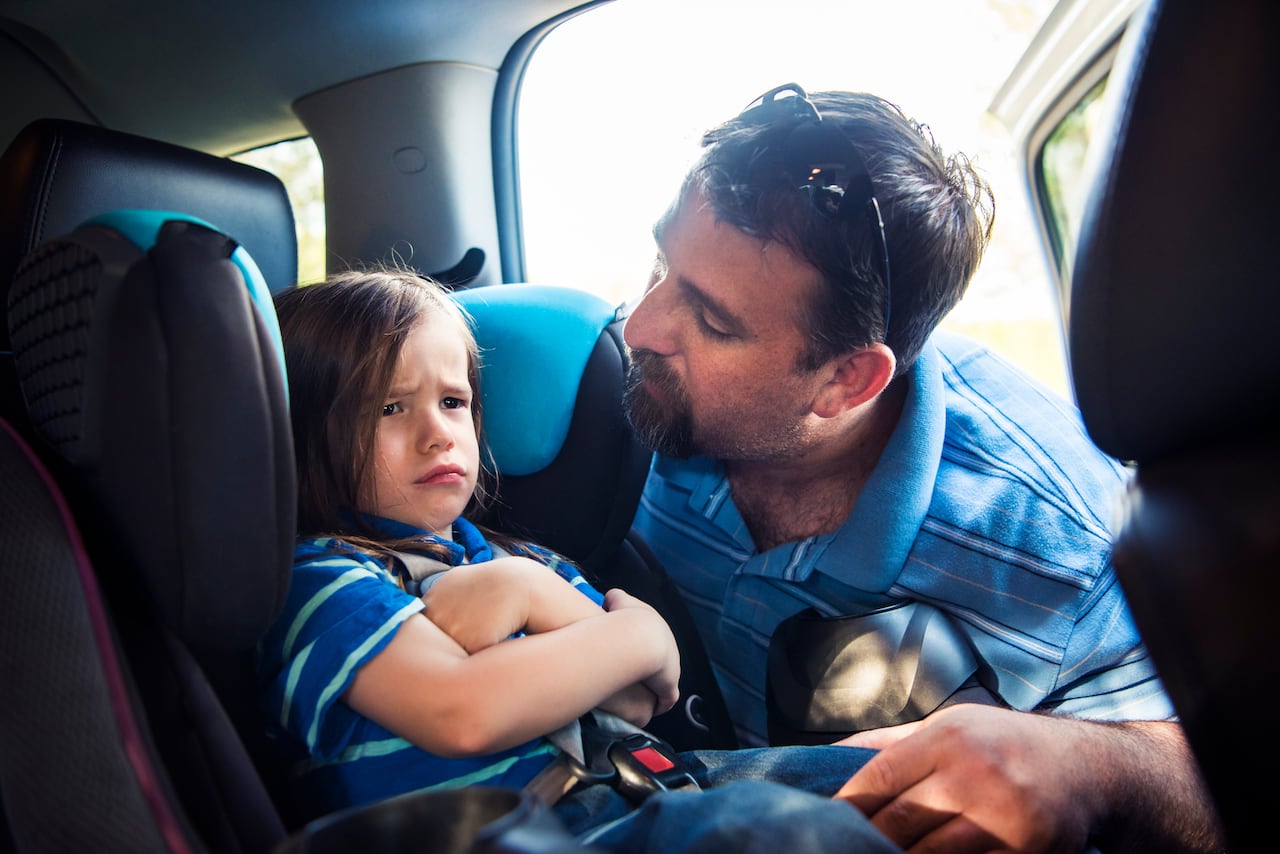 A father looks at his toddler son who is pouting in the car seat. 