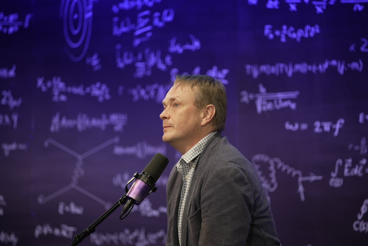 Mężczyzna ubrany w ciemnoszary garnitur siedzi przed mikrofonem podcastu.
