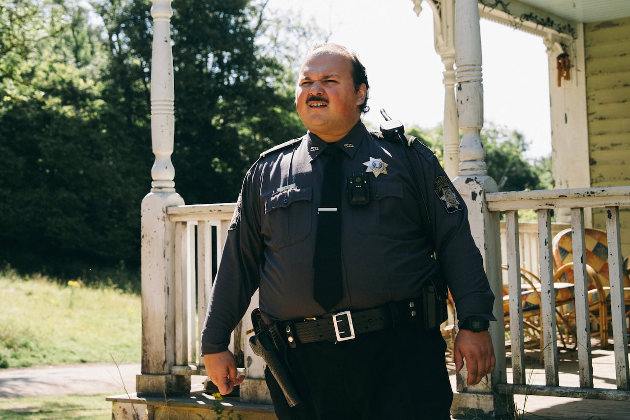 A man in a police uniform stands near a dilapidated porch.