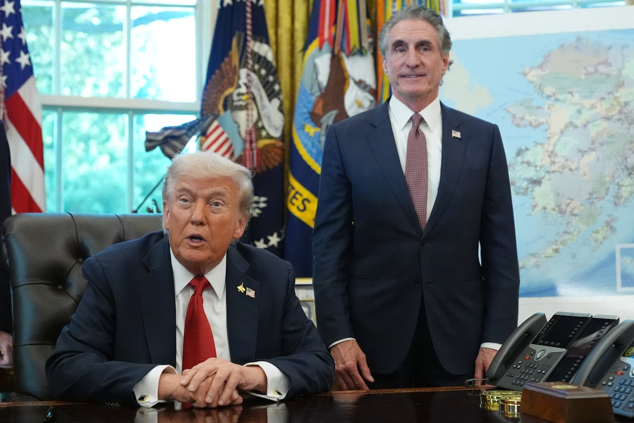 Donald Trump at his desk in the White House with a man in a suit standing behind him.
