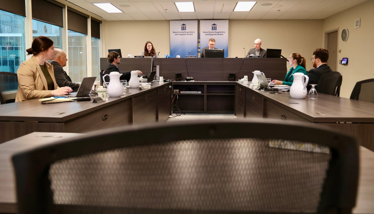 Men and women in suits and blazers sit at desks across from each other, below a higher desk where a woman and two men sit under banners
