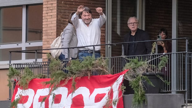 A man gestures from a balcony, from which hangs a large red banner that reads "REPORT" in block white text.