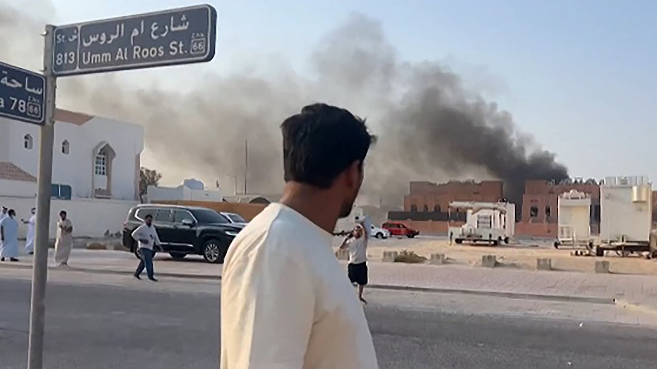 Man stands beside a street sign looking at a low-rise building with smoke billowing from it, and a handful of other people walking in the distance.