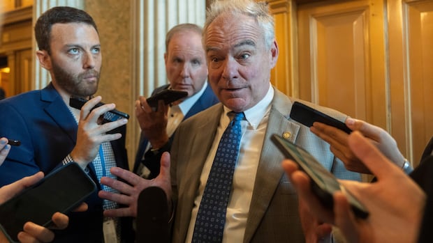 A man in a suit speaks to a group of reporters standing around him holding their phones. 