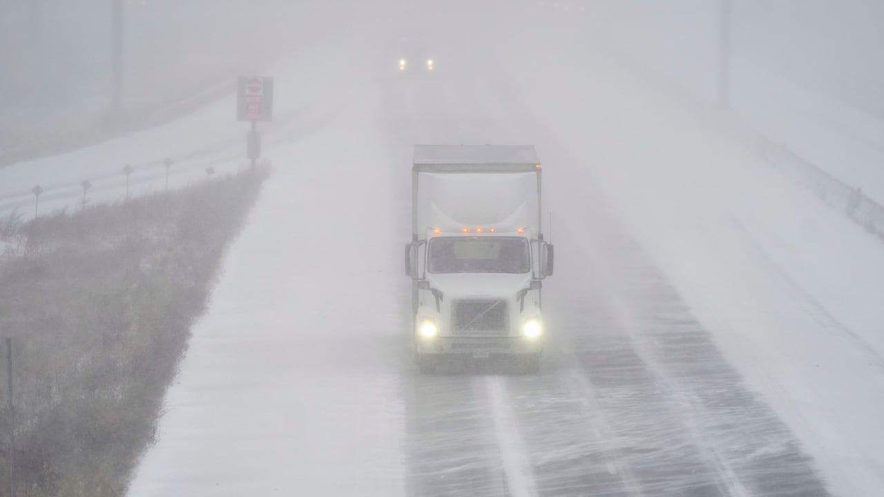 A large white truck drives along a snowy road. It is difficult to make out the vehicle behind the truck due to the blowing snow.