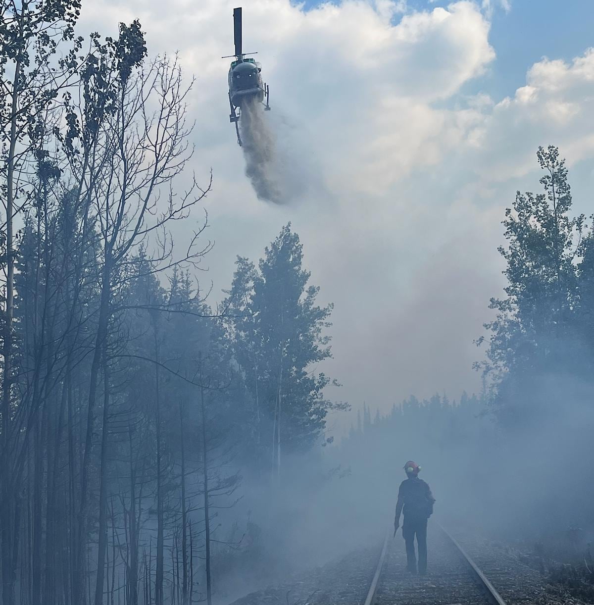 A firefighter, shrouded in smoke, looks on as a helicopter sprays water above him.