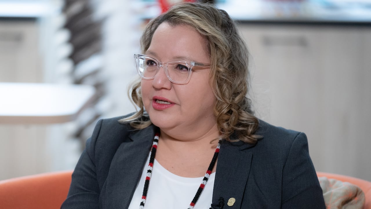 A woman with shoulder-length, dirty blonde hair, clear-rimmed glasses is pictured speaking in a sit-down interview.