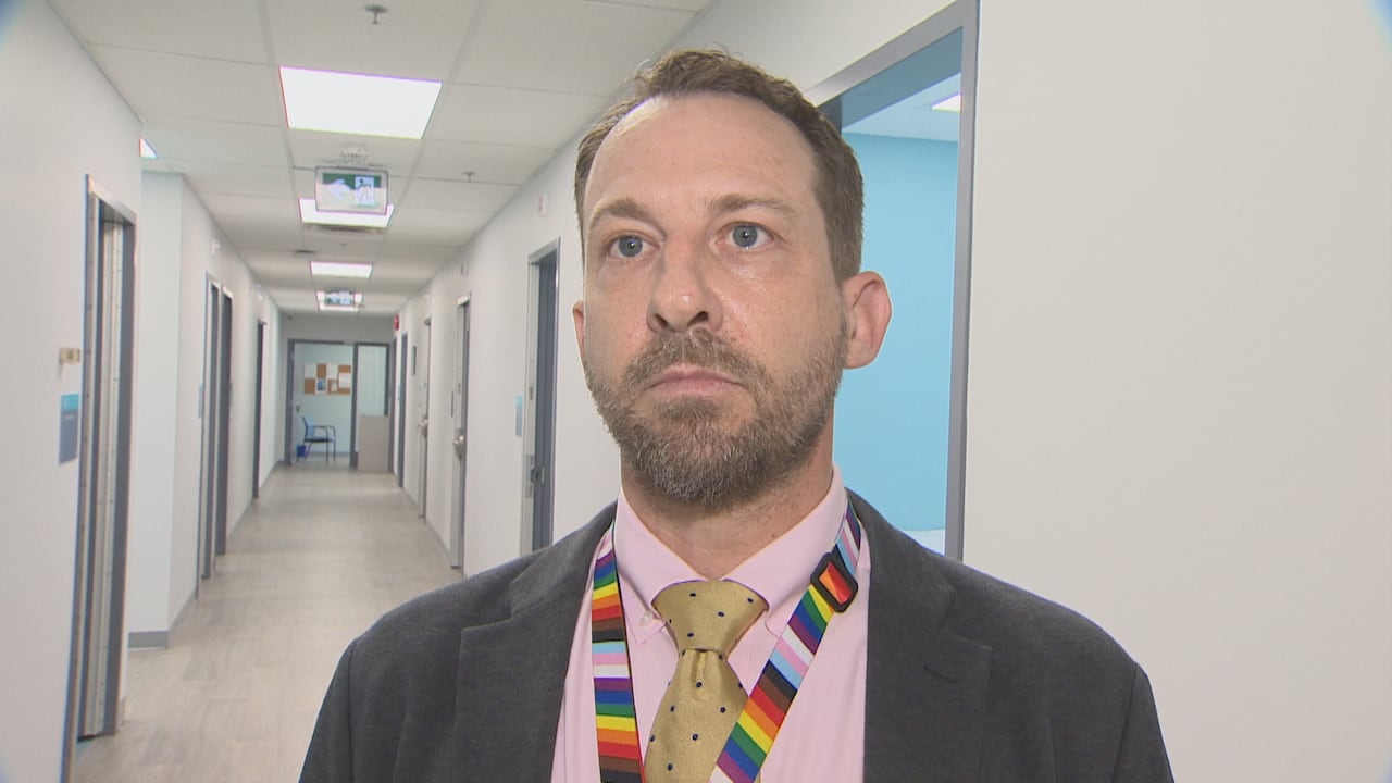 A antheral lasting successful a hallway looking off-camera. He is wearing a pinkish shirt, golden tie, grey suit jacket, with a rainbow lanyard astir his neck.