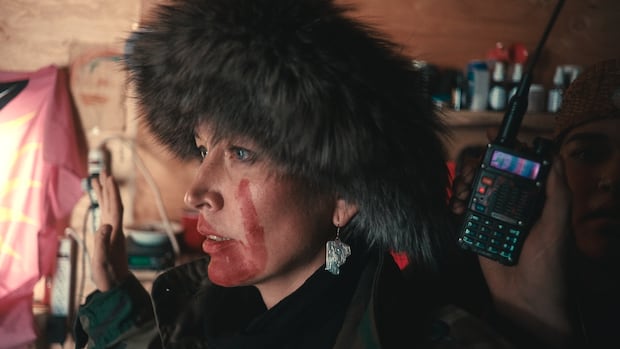 Tsakë ze' Sleydo' Molly Wickham holds up her hands as RCMP point guns at her during an arrest at Coyote Camp on traditional Gidimt'en territory on Nov. 19, 2021. Inside a cabin, a woman with red handprints painted on her face stands with her hands in the air, staring straight ahead. A hand holds up a walkie-talkie beside her.