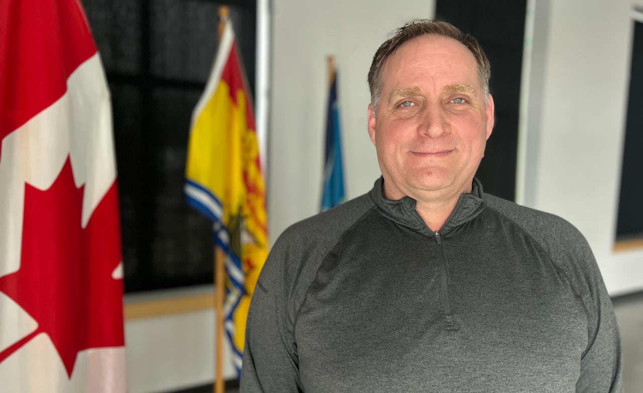 A antheral   with a grey sweater smiles astatine  the camera portion    lasting  successful  beforehand   of the Canadian and New Brunswick flags wrong  a bare room. 