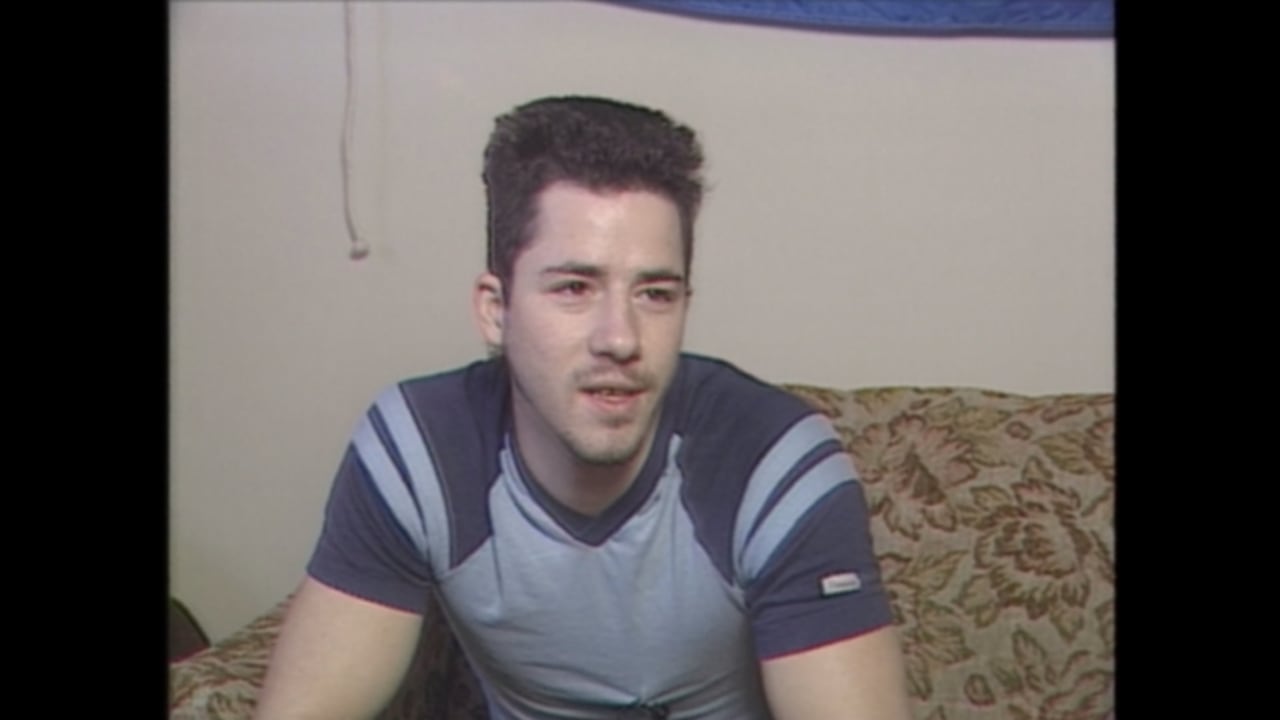 A man in a blue shirt with black hair sits on a brown patterned couch.