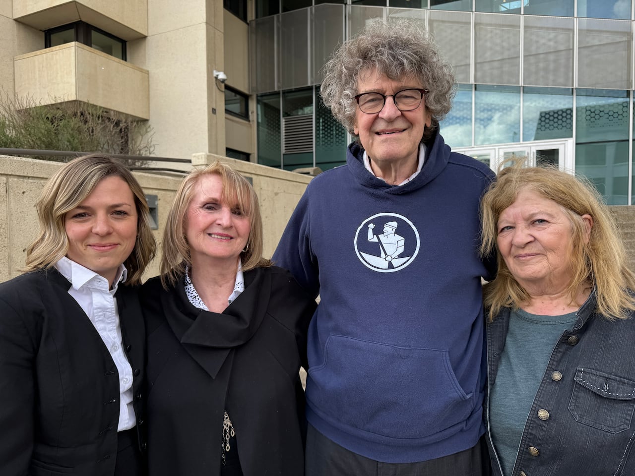 A man with grey curly hair stands outside a building, with two women, one wearing lawyer robes, on his left, and another woman on his right.