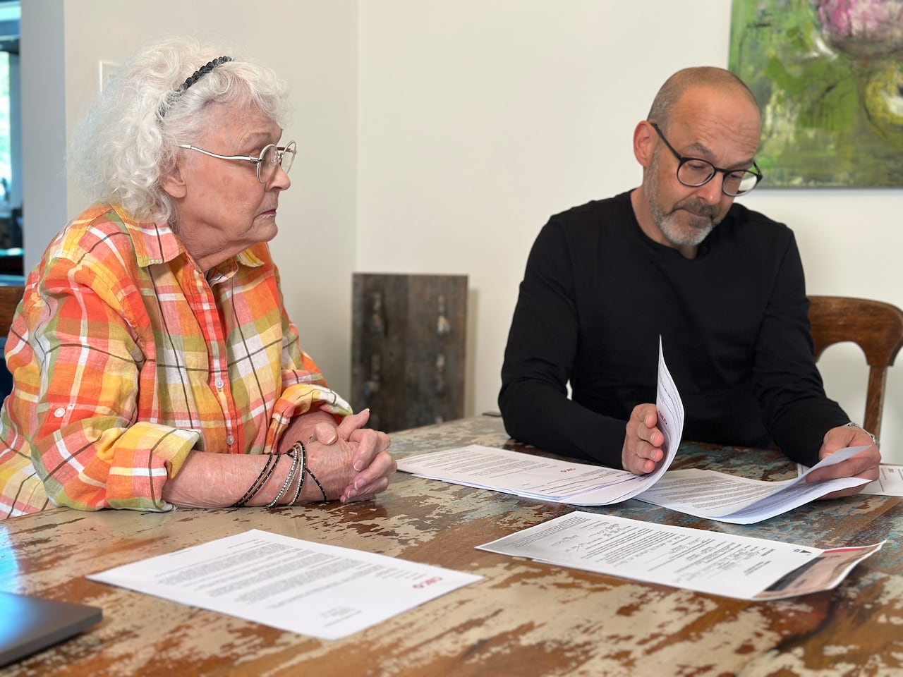 An older woman with curly white hair and glasses sits at a dining room table next to a balding middle-aged man with glasses as he flips through several pieces of paperwork. 