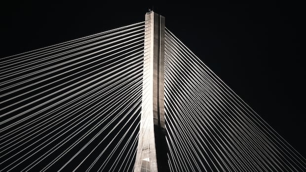 Cables on the Gordie Howe Bridge illuminated by the bridge's esthetic lights. Close-up of the top of one of of the bridge towers and the cables stretching from each side of it, all illuminated against the night sky. Nothing else is visible, making the geometry of the image look strikingly artistic.