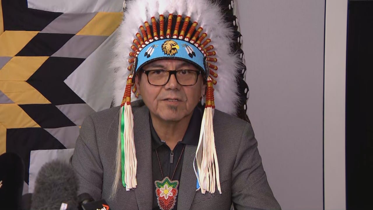 A man in glasses and wearing a headdress speaks at a microphone