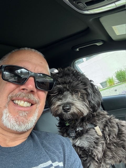 A smiling antheral   poses for a selfie adjacent  to a achromatic  dog.