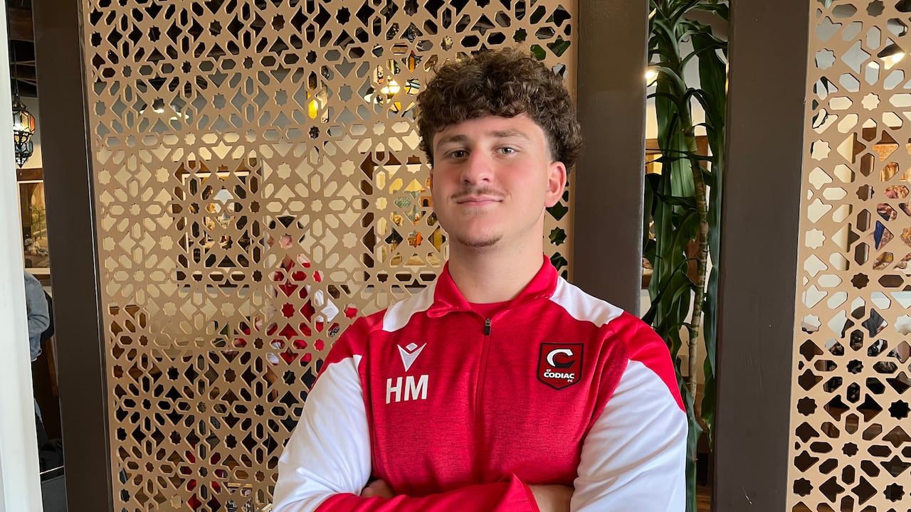 A young antheral   with abbreviated  curly hairsbreadth  poses with his arms crossed. He's wearing a work   and achromatic  agelong  sleeved jersey with the initials HM connected  his chest.