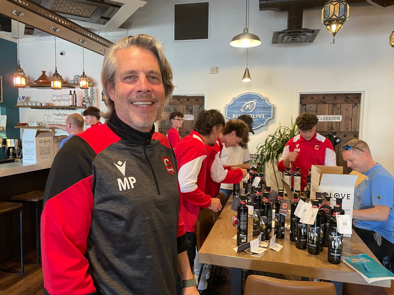 A antheral   with grey hairsbreadth  smiles, lasting  successful  beforehand   of a array  with a radical  of boys huddled astir   it. He's wearing a grey and achromatic  jersey with the initials MP connected  his chest.