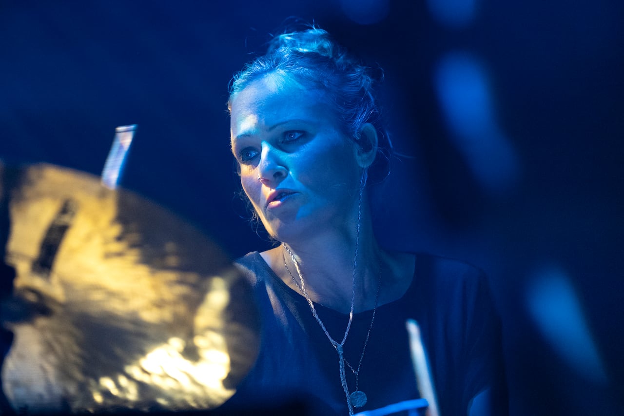 A woman with light hair is shown on a blue-lit state with her hair tied back playing the drums.