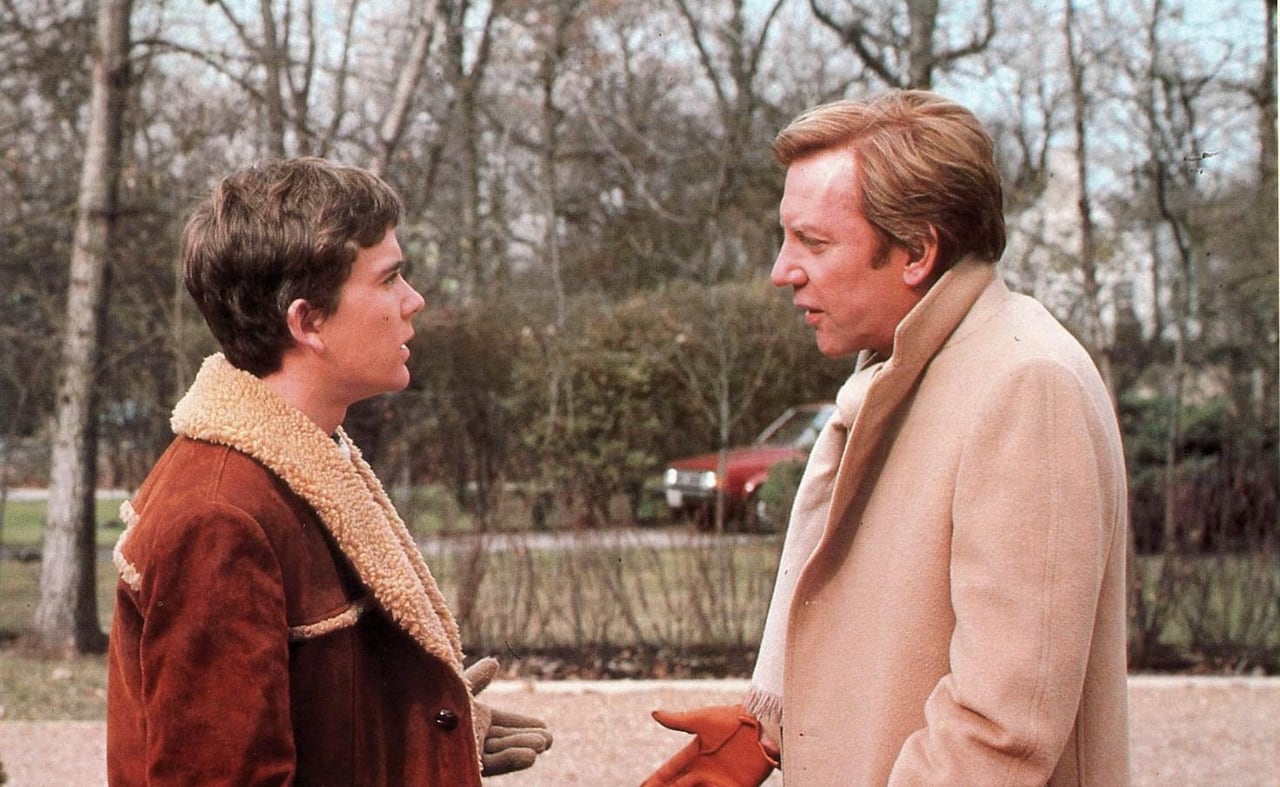 2 people talking in a driveway. They're wearing fall coats and gloves, and the leaves are off the trees behind them. 