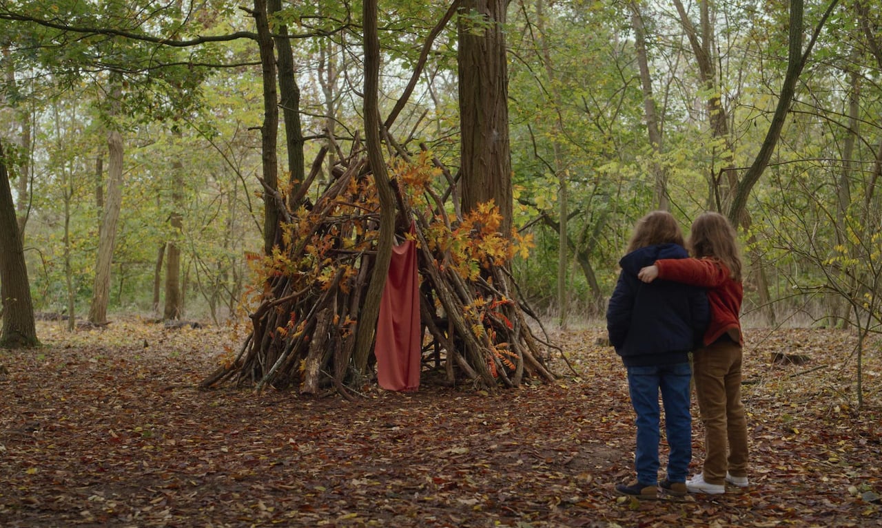 2 kids standing in a forest, looking at a fort made of sticks with lots of fall foliage attached to it. 