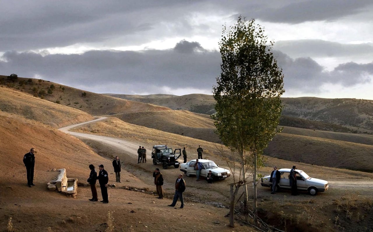 A group of men standing on the side of the road next to 3 cars, on a barren road among hills. 