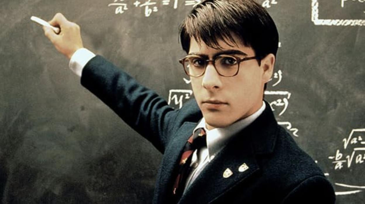 A man in a blazer and tie writing math on a chalkboard. 