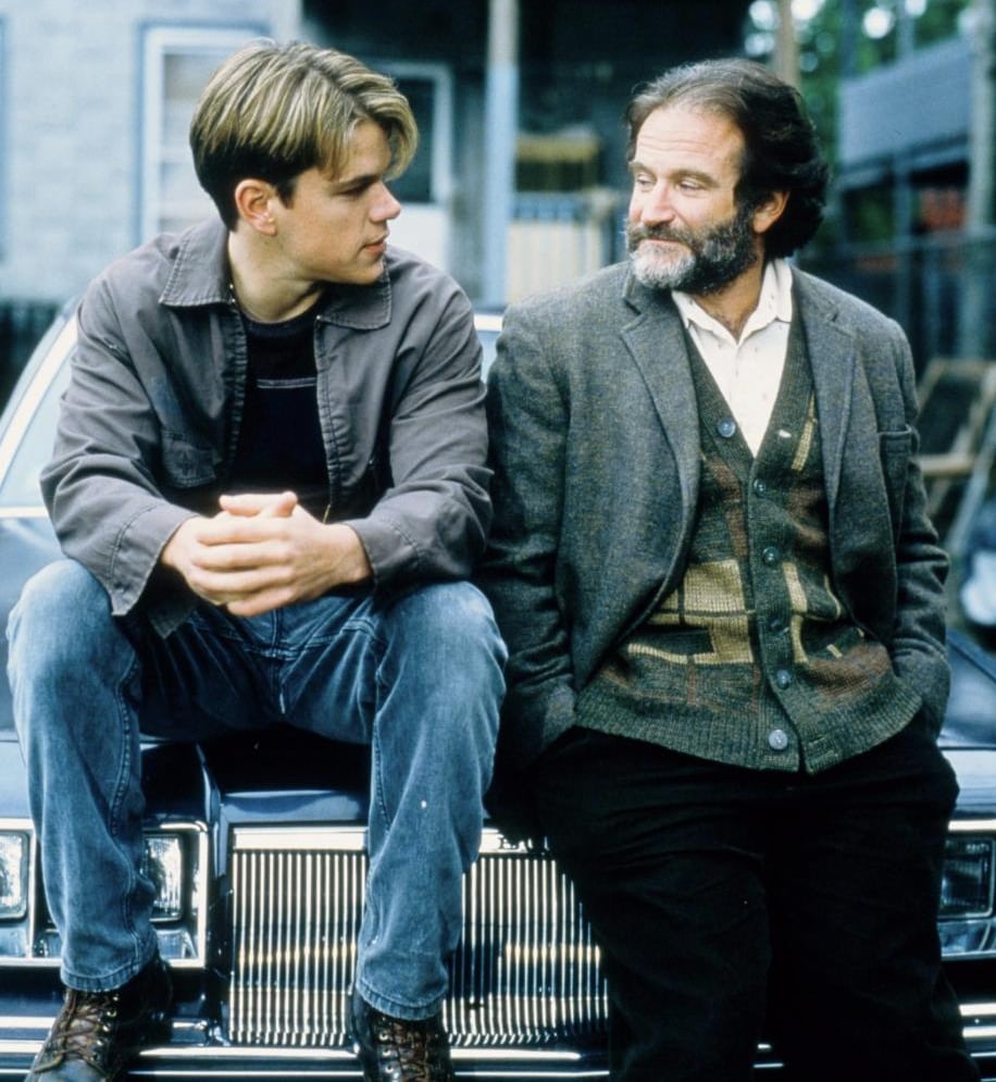 2 men sitting on the hood of a car in front of an apartment building.