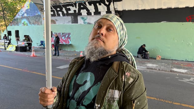 A man wearing a scarf and holding a pole wears a jacket covered in paint splatters.