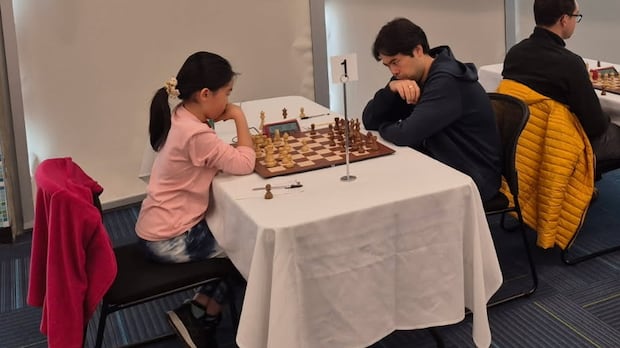 Two people play chess. One is a 12-year-old girl, wearing a pink sweater, and the other is a adult man, wearing a blue sweater