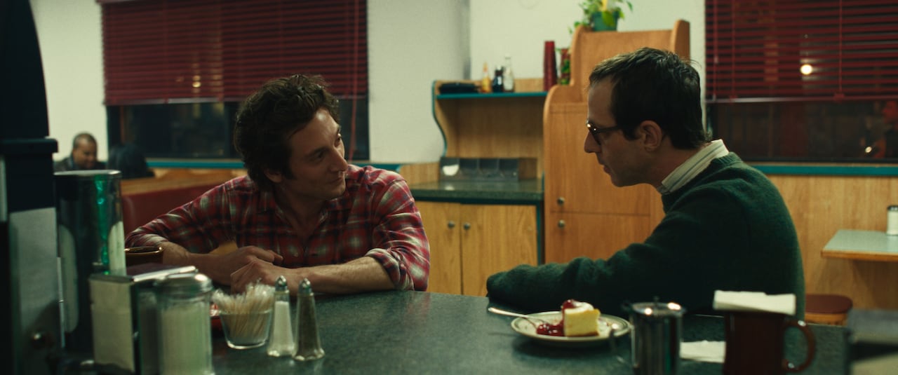 Two men sit at a diner counter. The man on the left is smiling wryly while the man on the right appears concerned.