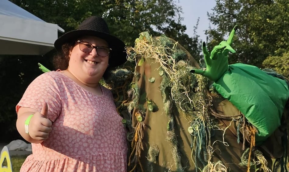 A pistillate successful a pinkish formal stands adjacent to a idiosyncratic wearing a greenish outfit resembling a swamp.