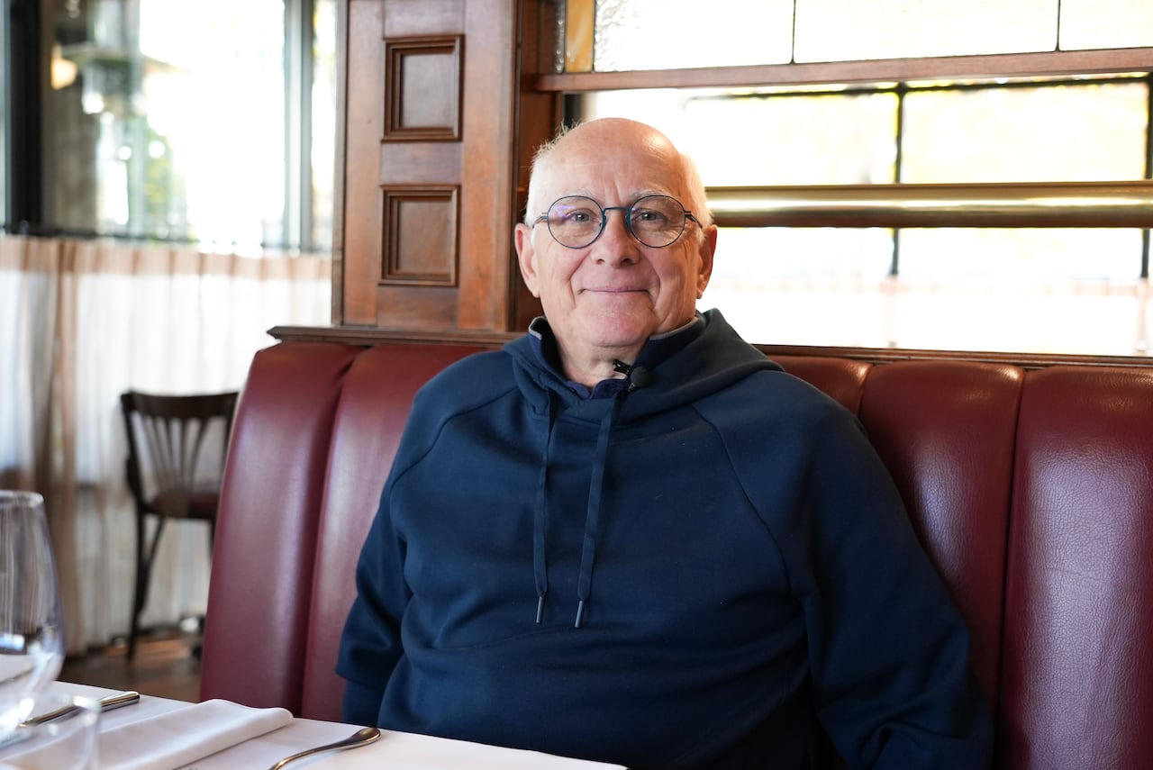 A gray-haired man in glasses smiles while sitting in a restaurant booth.