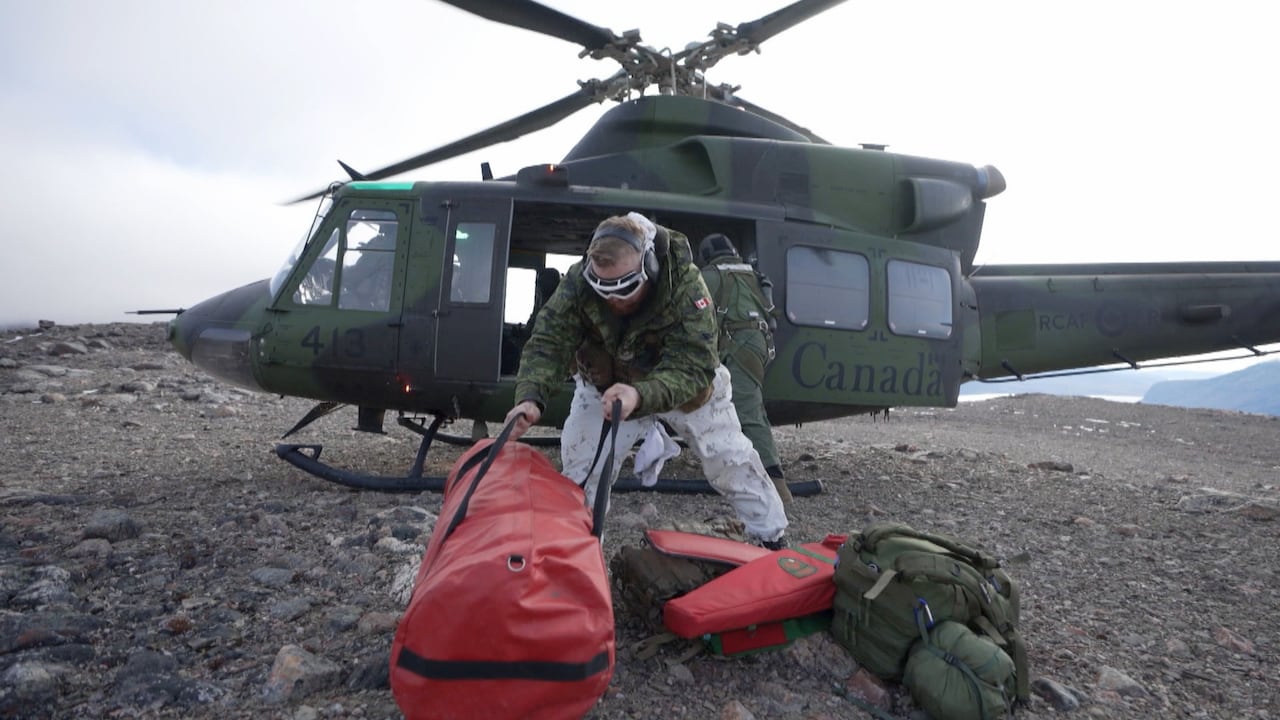 Man in goggles and camoflage coat drops red duffle bag in front of helicopter