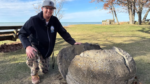 Man kneels beside a large round stone, lake in the background.