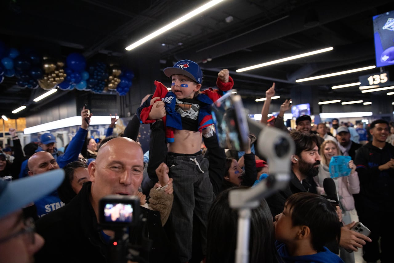 A man holds up a toddler decked out in Toronto Blue Jays clothing and make up as a crowd inside an arena celebrates.