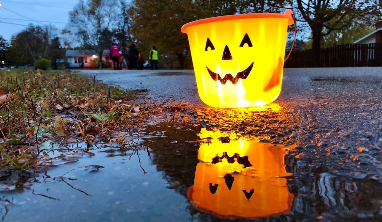 A glowing jack-o'-lantern basket sits on a wet sidewalk and his face is reflected in a puddle.