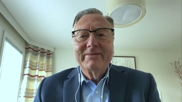 A person in a blue blazer and glasses smiles gently while on a video call. Light curtains and framed artwork are visible in the bright room.
