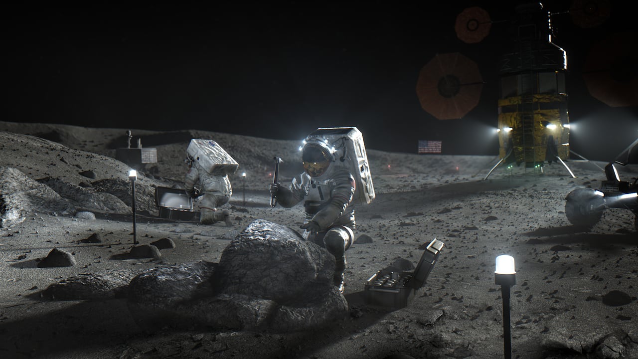 A person takes a hammer to a rock on the moon, while another person is in the background along with other tools and a spacecraft.