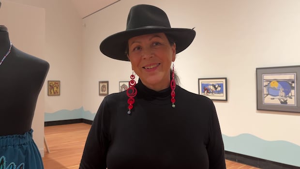 Kym Gouchie is the co-curator of the Too Beh Ts’ughuna: Water is Life exhibit at the Two Rivers Gallery in Prince George A woman in a hat and earnings is smiling as she stands in an art gallery.