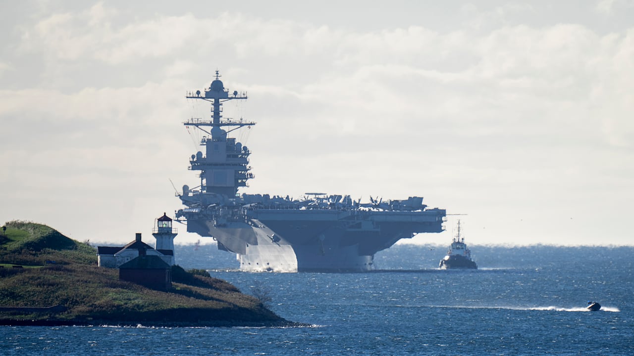 A large aircraft carrier sails near a coastline.