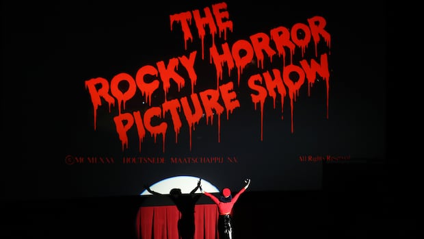 Happy To Be Here cast member performs during the screening of "The Rocky Horror Picture Show" during the 2025 TCM Classic Film Festival at TCL Chinese Theatre on April 25, 2025 in Hollywood, California. A woman in a red and black outfit stands in a spotlight looking up at a large screen that reads "The Rocky Horror Picture Show."