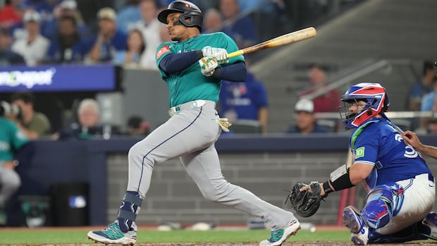 A baseball player in a turquoise jersey swings a bat as a catcher in a blue jersey and blue, white and red helmet squats behind him.