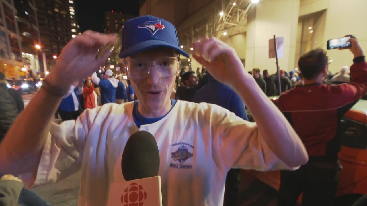 A young man in a Jays ballcap and white sweatshirt with a crest commemorating the Toronto Blue Jays 1992 World Series win, stands in front of a crowd of fans in downtown Toronto at night