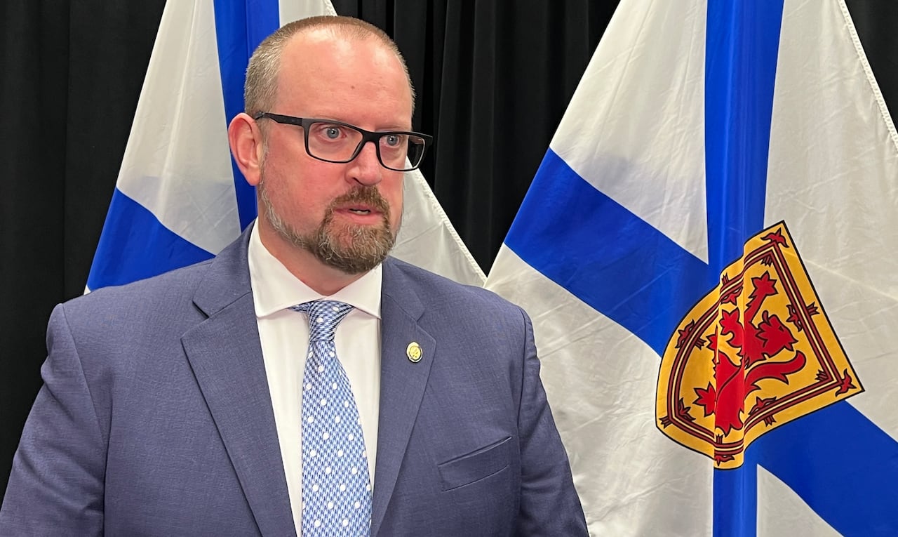 A antheral successful a suit and necktie with glasses stands by a Nova Scotia flag.