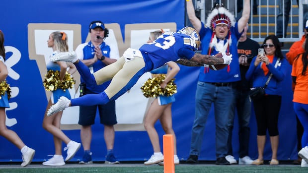 A ball carrier in blue and gold stretches his arms out as he flies through the air into the end zone.