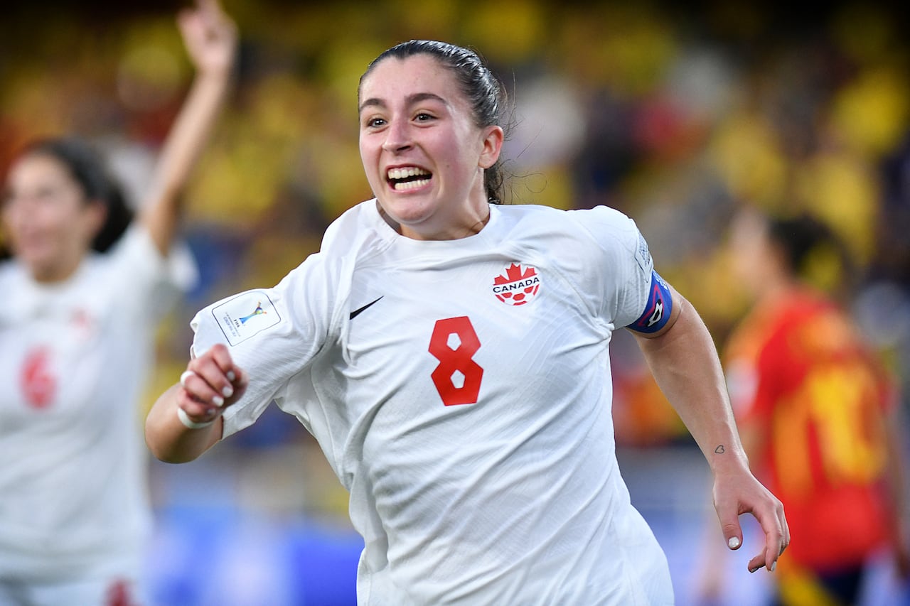 A female soccer player in a white Canada jersey celebrates after scoring a goal.