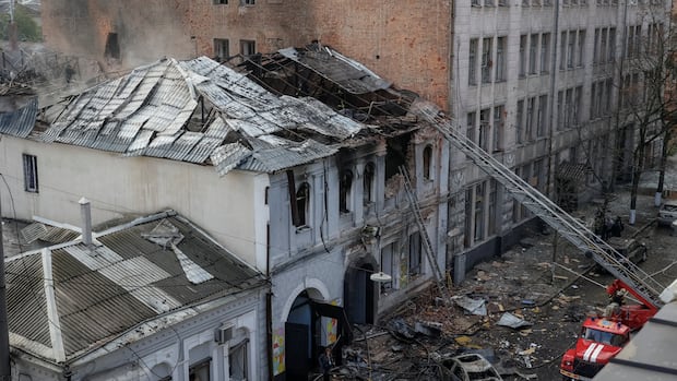 Destroyed building with a collapsed roof, debris scattered on the ground, and a damaged car. Fire truck ladder extends to the upper floor, conveying a scene of devastation.