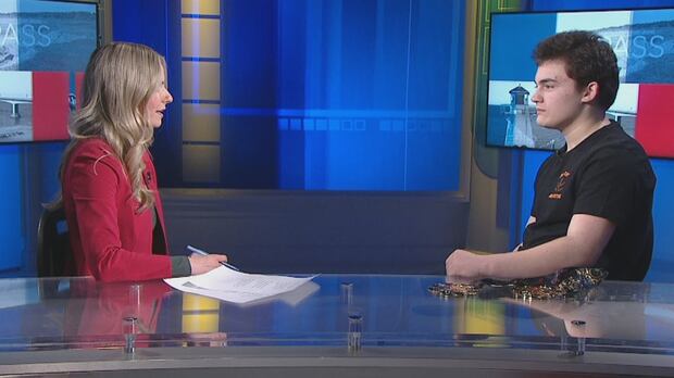 Thomas Dumas talks to Sheehan Desjardins as she hosts CBC News: Compass. A blonde woman in a red blazer talks to a teenager with dark hair who's wearing a black T-shift and has several medals on the desk next to him.
