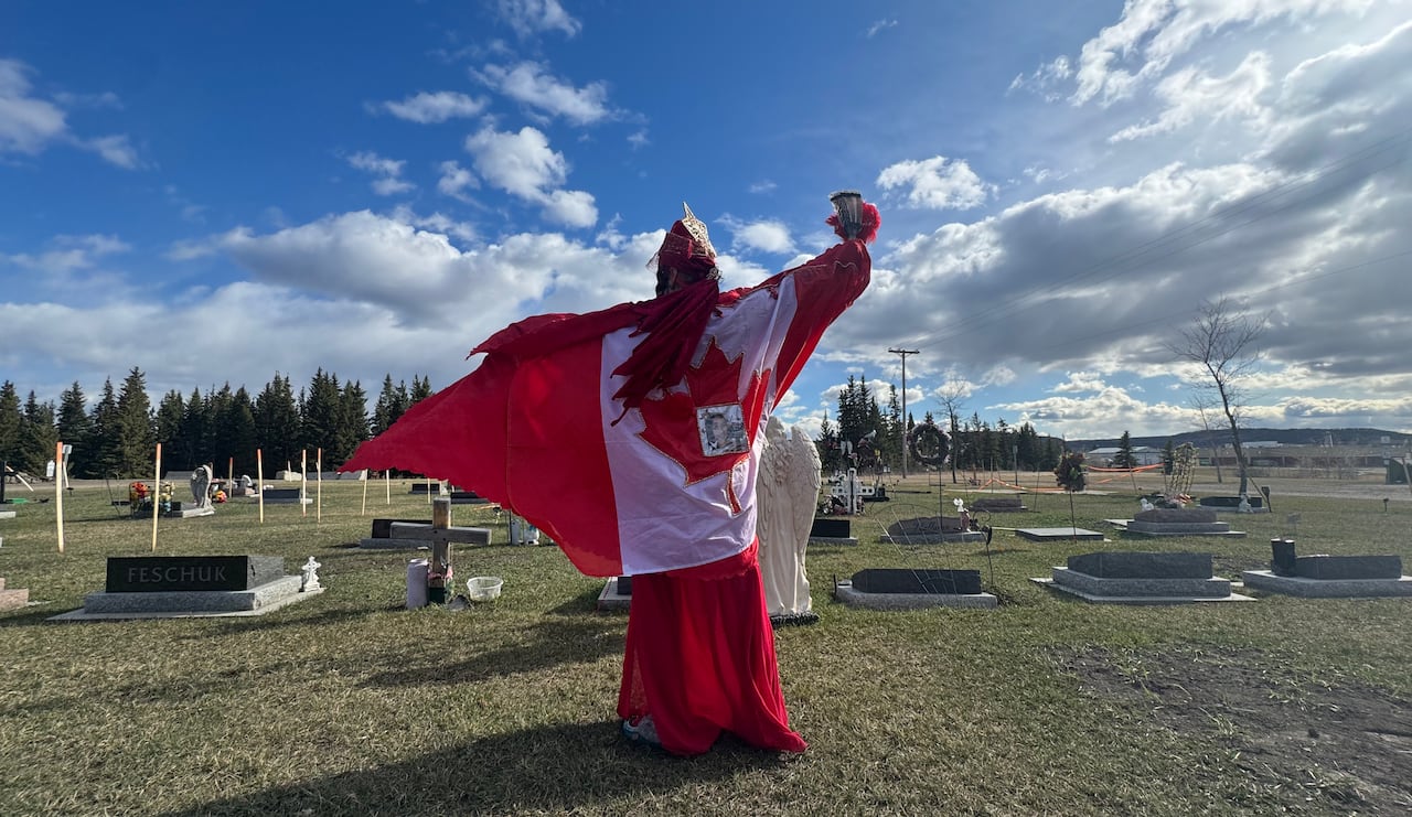 A cree woman in regalia holds up her arm while standing in a cemetery. She is draped in the Canadian flag.